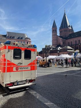 Man sieht links die Rückseite eines Malteser Rettungswagens und auf der rechten Seite den Dom von Mainz sowie den Markplatz