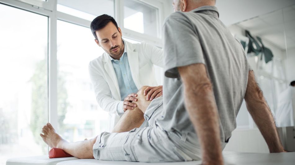 Ein Arzt untersucht das Knie eines Patienten, der auf einer Behandlungsliege sitzt.
