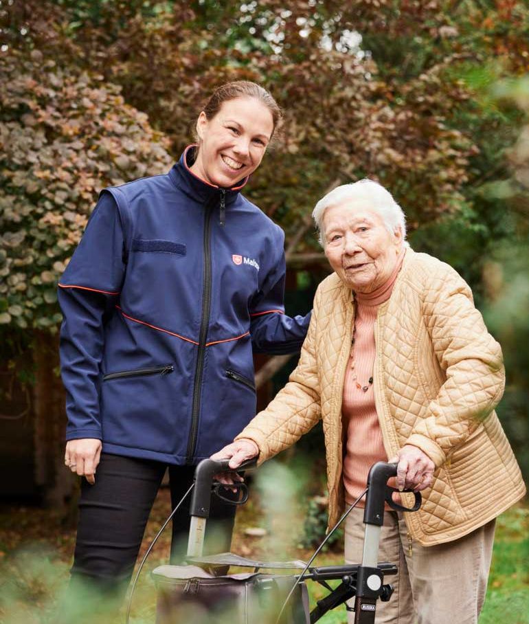 Eine junge Frau in Malteser Jacke und eine ältere Dame mit Rollator stehen im Grünen und lächeln.