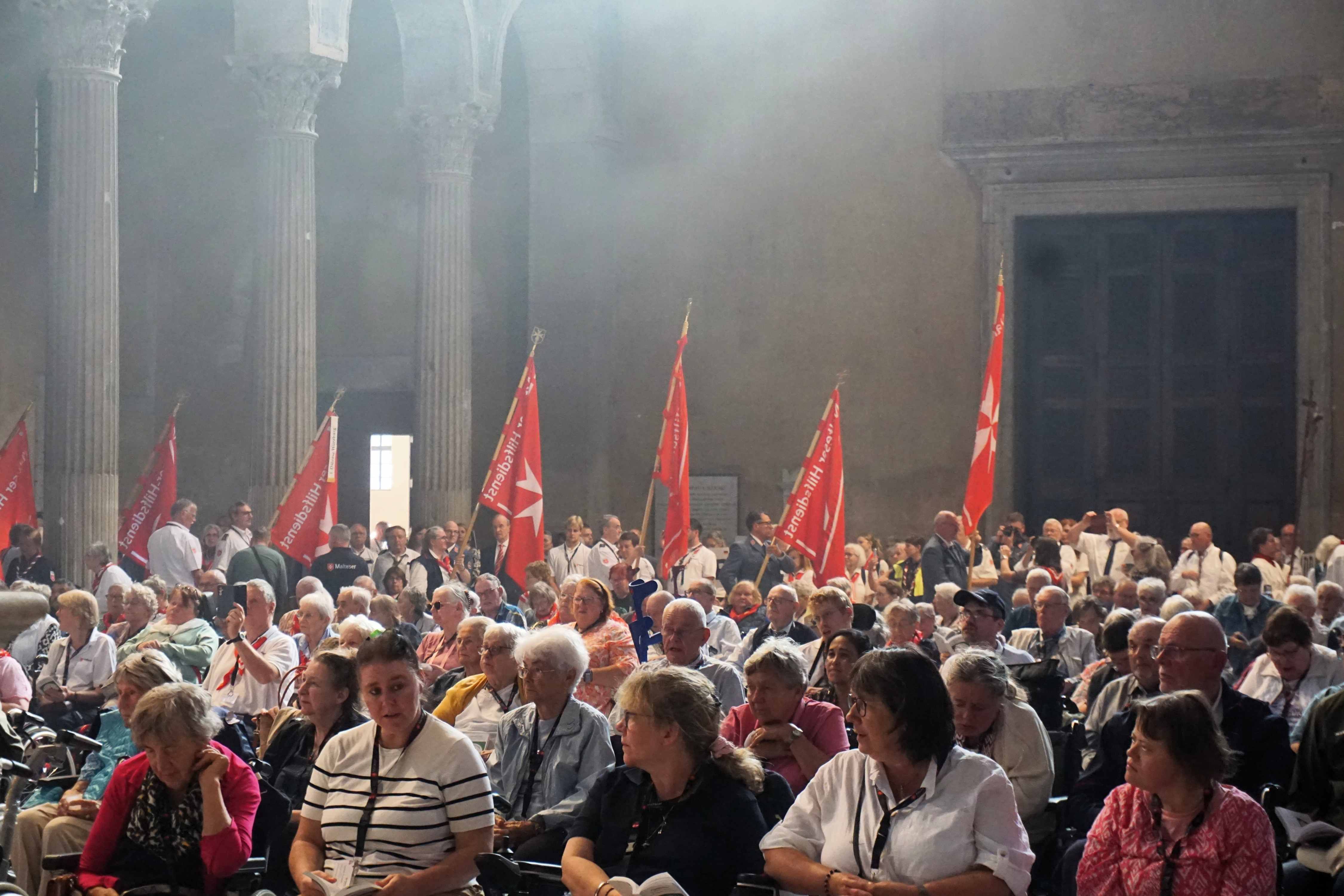 Große Menschenmenge in einer Kirche, viele halten rote Fahnen, helle Lichtquelle von oben, hohe Säulen im Hintergrund.