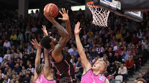 Spieler der Bamberg Baskets beim Korbleger gegen zwei Verteidiger in Pink vor vollen Zuschauerrängen.