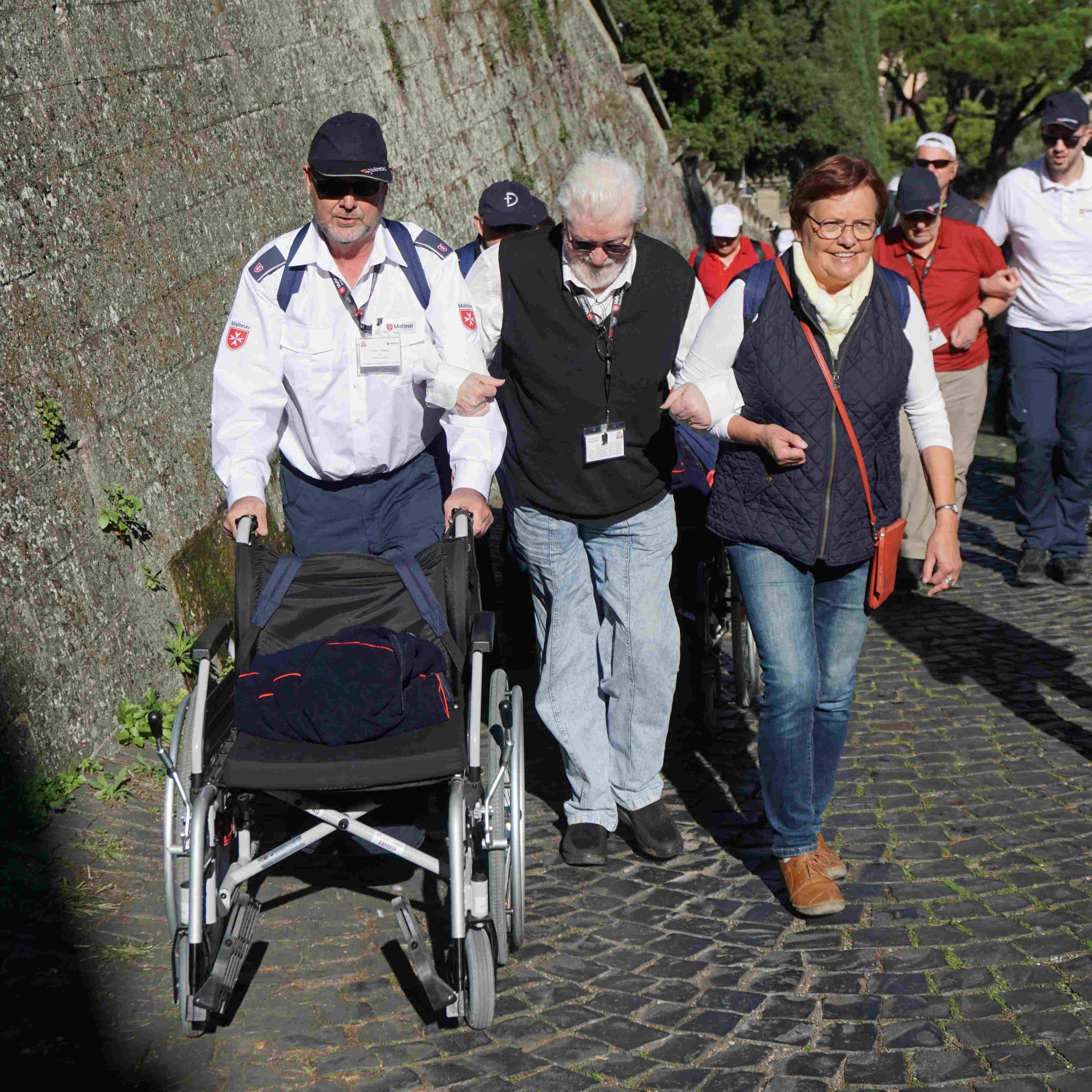 Drei Personen gehen auf einem gepflasterten Weg in Castel Gandolfo, eine schiebt einen leeren Rollstuhl, umgeben von weiteren Spaziergängern.