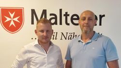 Zwei Männer stehen vor einer Wand mit Malteser Logo