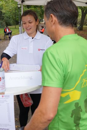 Eine ehrenamtliche Mitarbeiterin der Malteser begrüßt einen Läufer am Stand und weist ihn ein.