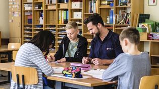 Drei Kinder sitzen am Tisch und werden von einem Helfer der Malteser unterstützt