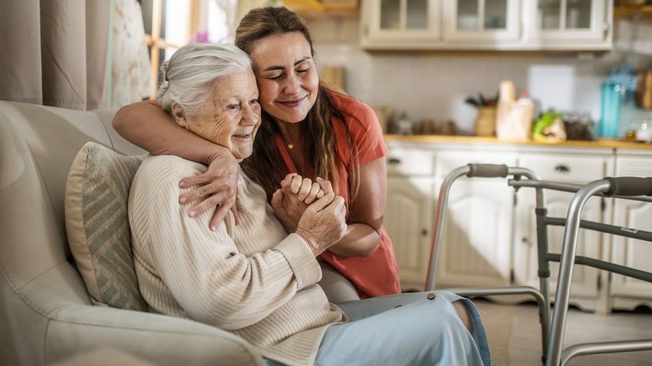 Eine junge Frau legt lächelnd den Arm um eine ältere lächelnde Frau auf einem Sofa, hinter dem eine Küchenzeile zu sehen ist.
