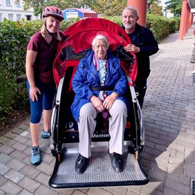 Eine Frau sitzt in einer Fahrradrikscha. Neben ihr stehen eine Frau und ein Mann.