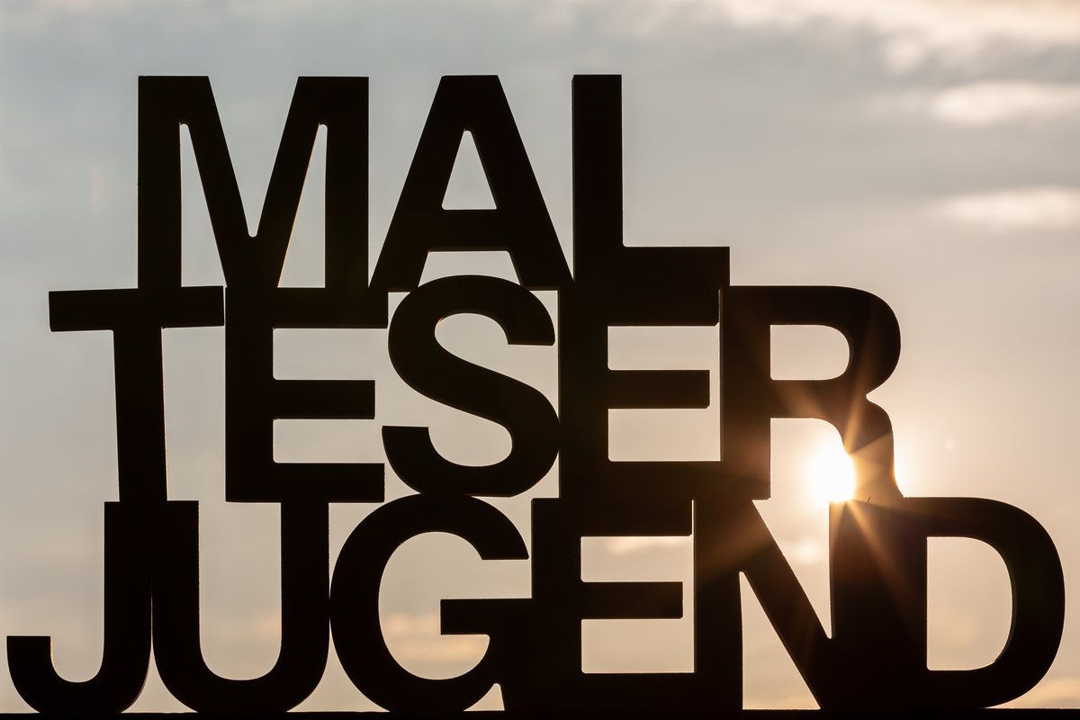 Das Bild zeigt große, fettgedruckte schwarze Buchstaben in gestapelter Anordnung vor einem Hintergrund aus bewölktem Himmel mit teilweise sichtbarer Sonne. Die Buchstaben formen den Schriftzug „MALTESER JUGEND“.