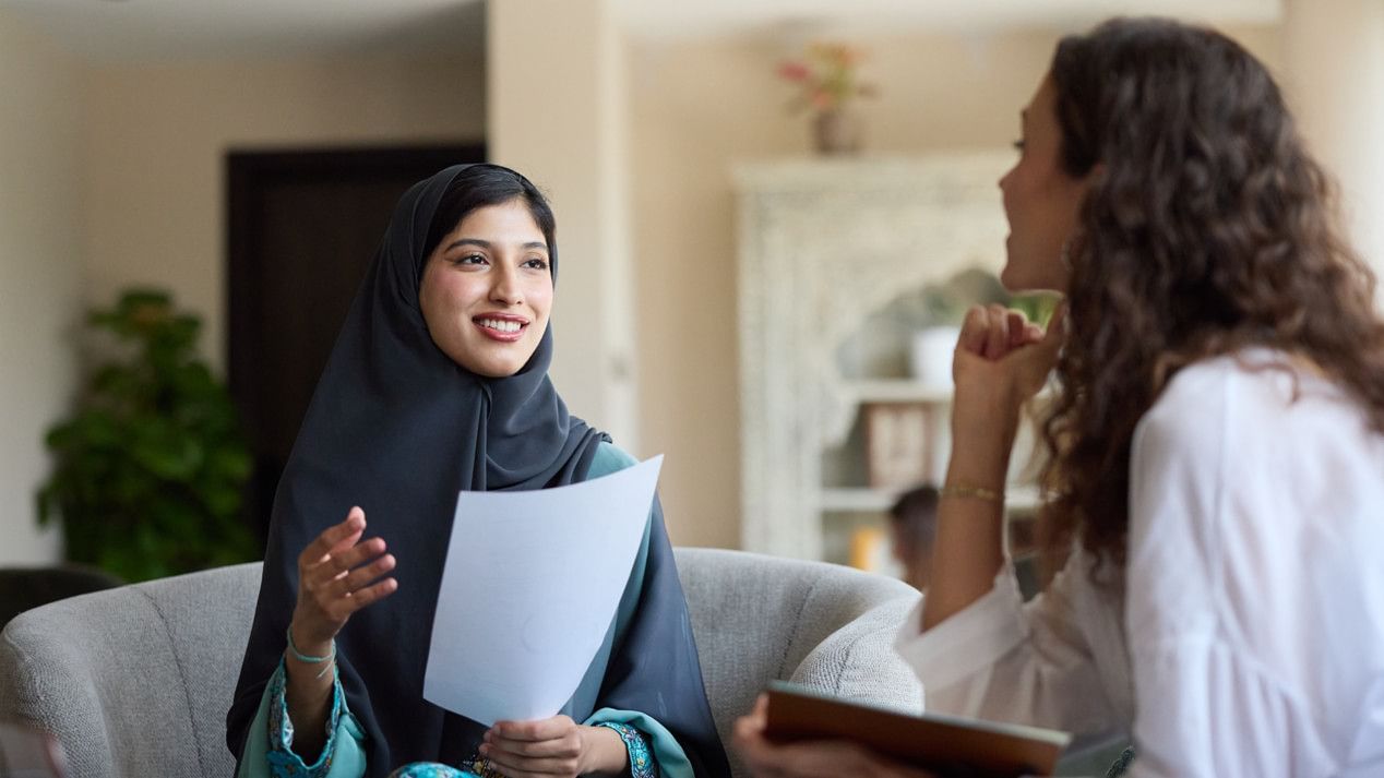 Eine lächelnde junge Frau mit Hijab hält ein Blatt Papier in der Hand und unterhält sich mit einer weiteren jungen Frau.