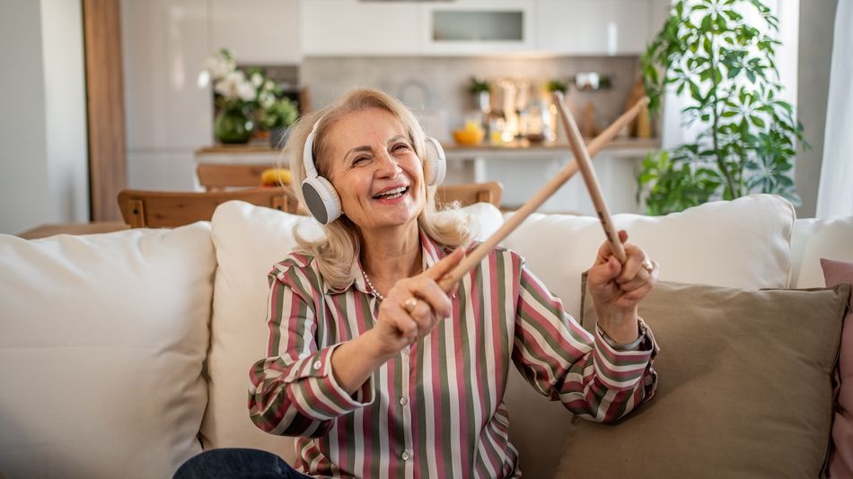 Eine ältere Frau mit Kopfhörern und Drumsticks sitzt lächelnd auf einem Sofa