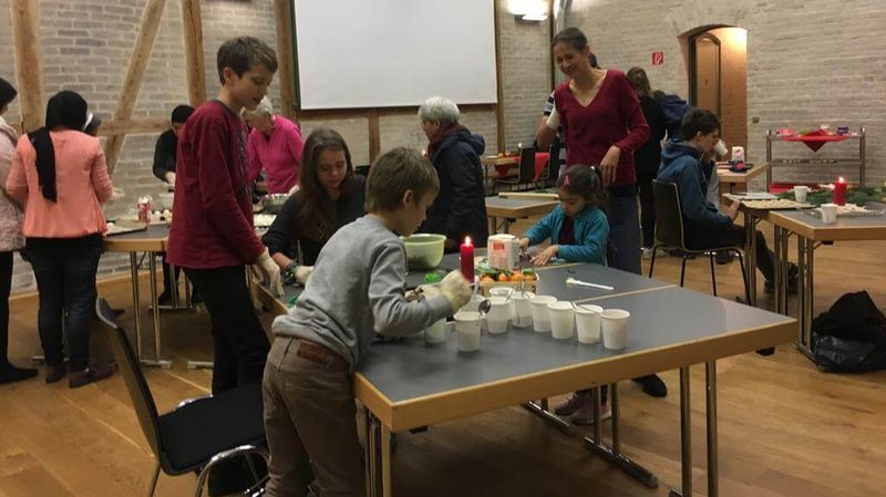 Kinder und Erwachsene in einer Halle mit Tischen, auf denen weihnachtliche Backutensilien stehen.