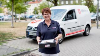 Mitarbeiterin des Malteser Menueservice trägt eine Warmhaltebox und lächelt in die Kamera.