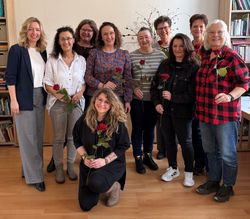 Eine Gruppe von Personen steht in einem Raum vor einer Bücherwand zusammen und hält jeweils eine einzelne rote Rose in der Hand. Eine Person kniet in der ersten Reihe, die übrigen stehen dahinter in zwei Reihen. Der Raum hat einen Holzfußboden, Regale mit Büchern und Ordnern sowie einen schlichten, hellen Hintergrund.