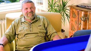 Mann sitzt auf gelbem Sessel in hellem Raum, im Vordergrund hält eine Person eine blaue Wäschewanne mit roter Kleidung.