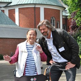 Eine Lachende ältere Dame und ein lachender Mann vor einer Basilika.