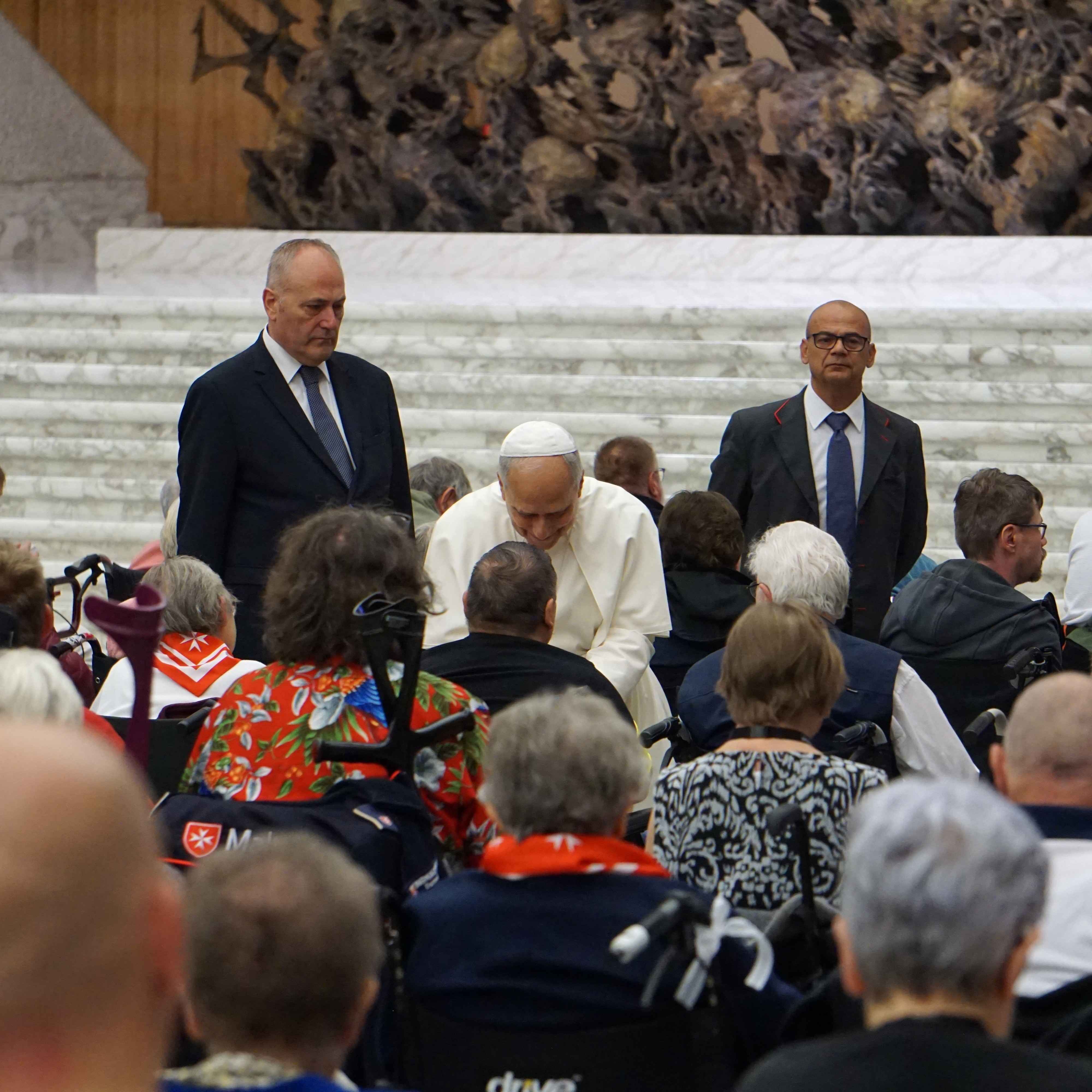 Person in weißer Kleidung sitzt vor einer Gruppe von Menschen, zwei Personen in Anzügen stehen seitlich, Hintergrund mit Stufen und Pflanzen.