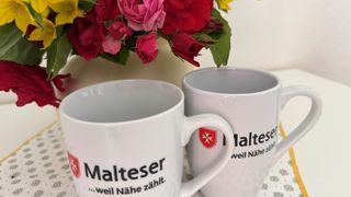 Zwei Tassen mit dem Logo und Claim der Malteser sind zu sehen, im Hintergrund ist ein schöner roter Blumenstrauß.