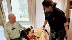 Ein Senior sitzt auf seinem Bett und streichelt Hund Finn, der neben ihm auf dem Bett liegt, daneben steht Frauchen Danielle von den Maltesern im Berchtesgadener Land.