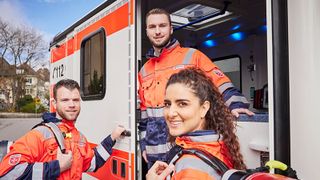 Mitarbeitende im Rettungsdienst in Einsatzkleidung stehen am Rettungswagen und lachen. 