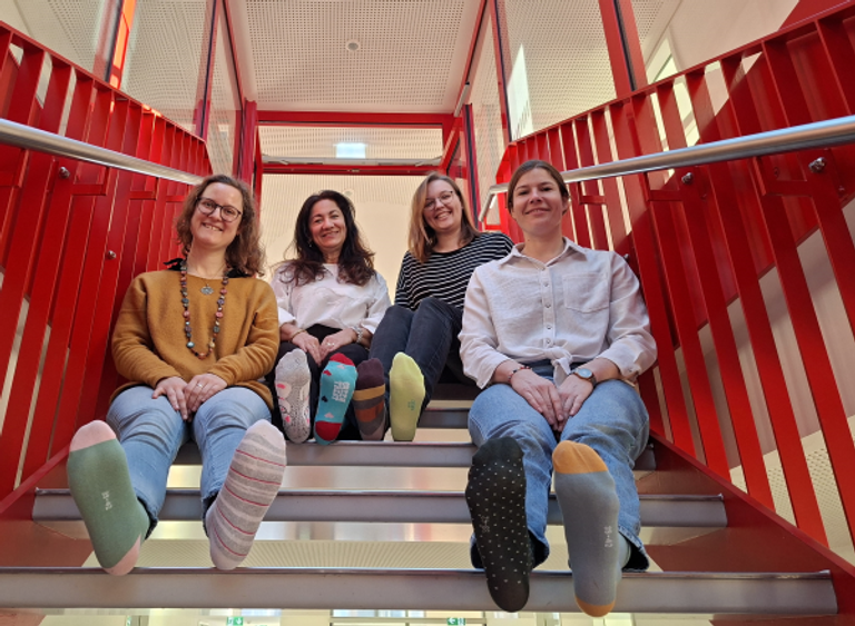 Vier Frauen sitzen auf einer Treppe und halten ihre mit unterschiedlichen bunten Socken bekleideten Füße in Richtung Kamera