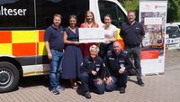Sieben Personen mit einem symbolischen Scheck stehen oder knien vor einem Krankenwagen