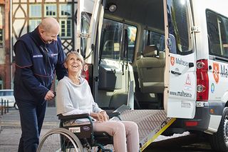 Malteser Fahrdienst. Eine gehbehinderte Frau im Rollstuhl wird vom Maltesermitarbeiter zum Fahrzeug geschoben.