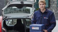 Ein junger Mann in Malteser-Kleidung steht mit einer Isolierbox in den Händen vor der geöffneten Heckklappe eines Autos.