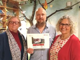 Zwei Frauen und ein Mann halten einen Scheck.