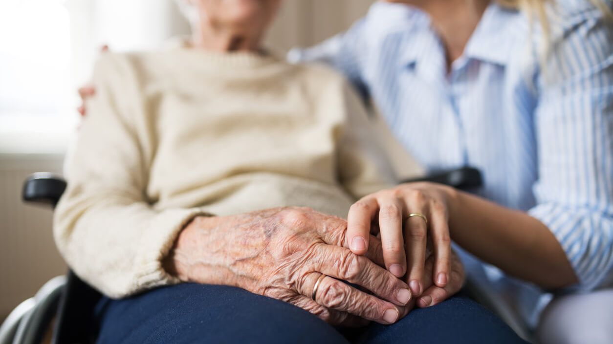 Eine ambulante Pflegekraft hält die Hand einer Seniorin im Rollstuhl.