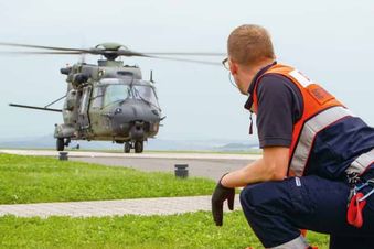 Malteser Helfer hockt auf dem Boden und schaut auf einen landenden Hubschrauber.