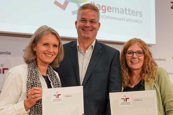 Drei Personen mit Zertifikaten bei einer Preisverleihung von „audit berufundfamilie“ stehen vor einer Leinwand.