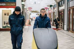 Ein Mann schiebt eine Renigungsmaschine durch ein Gebäude und ein anderer Mann läuft daneben.