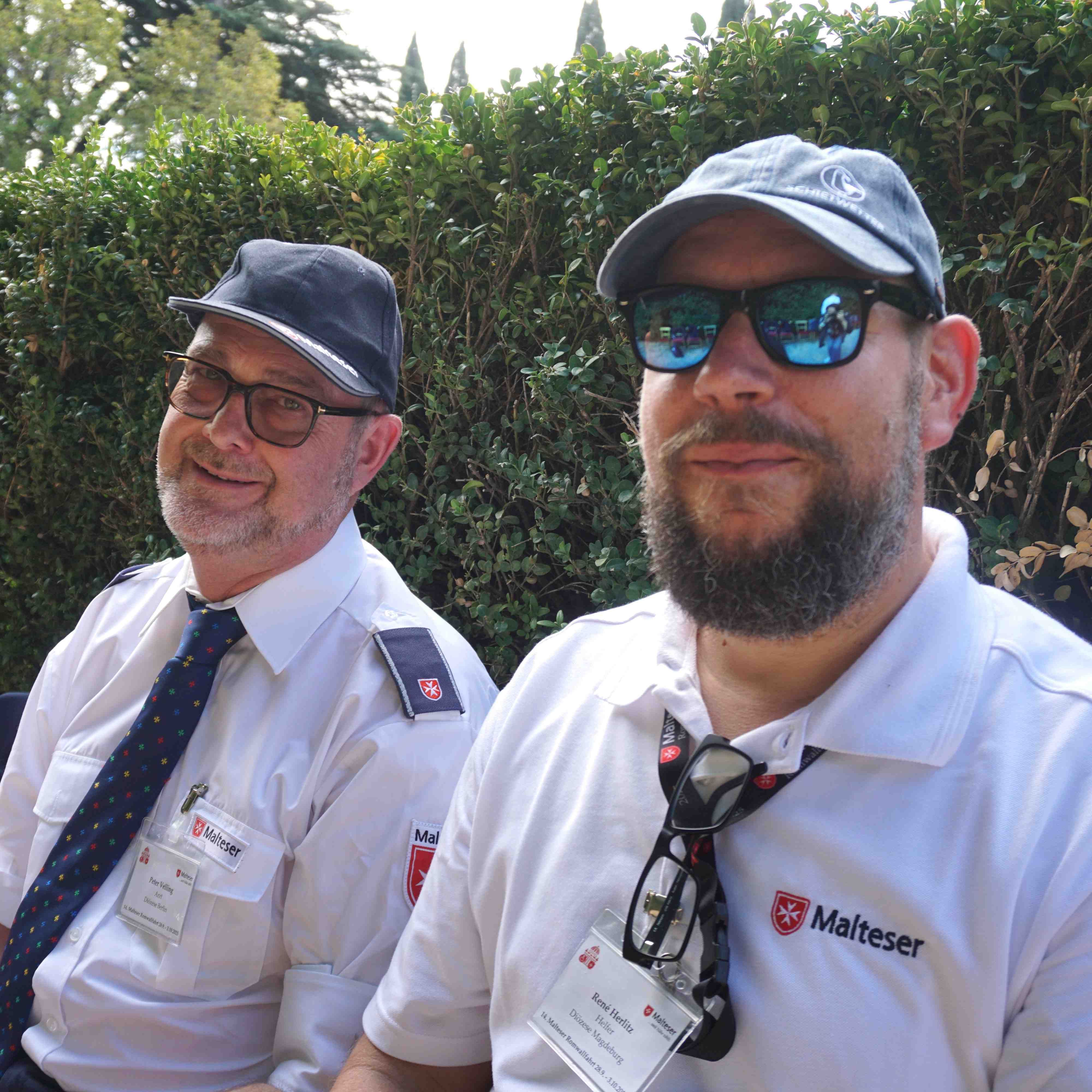 Zwei Männer in Uniform mit Kappen, einer trägt ein weißes Hemd mit Namensschild und Brille am Kragen, der andere ein weißes Hemd mit Krawatte.