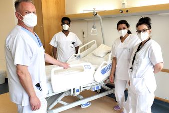 Vier Pflegekräfte in weißer Dienstkleidung und Maske an einem Klinikbett in einem Patientenzimmer.