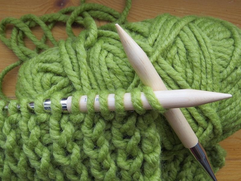 Gestricktes Stück in aus grüner wolle auf zwei überkreuz liegenden Strciknadeln