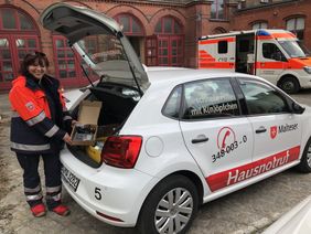 Malteser-Mitarbeiterin zeigt Hausnotruf-Gerät vor einem Einsatzfahrzeug der Malteser