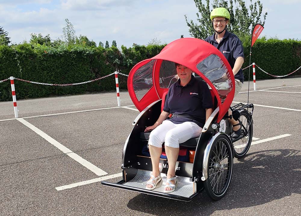 Eine Frau sitzt auf dem Gästesitz in einer Fahrradrikscha. Dahinter sitzt eine weitere Frau. Sie trägt einen Fahrradhelm.