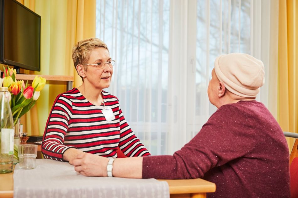 Eine ältere Frau beugt sich lächelnd über ein Sofa, in dem eine hochaltrige lächelnde Frau mit einer Decke sitzt.