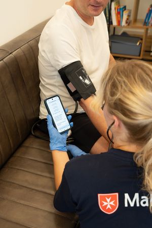 Das Bild zeigt eine Malteser-Mitarbeiterin, die einem Patienten den Blutdruck misst und die Daten in den TI-Messenger eingibt. 
