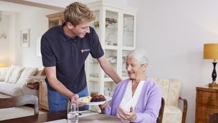 Ältere Frau in lilafarbenem Oberteil an einem Esstisch, junger Mann in Malteser Polohemd reicht ihr Essen auf einem Porzellanteller, 