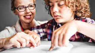 Frau sitzt hinter einem Jungen, der in Blindenschrift liest.