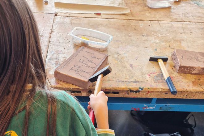 Eine Person sitzt an einer Werkbank und bearbeitet mit Werkzeugen eine Steinplatte. Auf dem Tisch liegen weitere Materialien und Arbeitsgeräte.