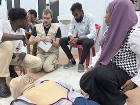 Eine Gruppe Menschen sitzt um eine Puppe. Foto: Malteser International