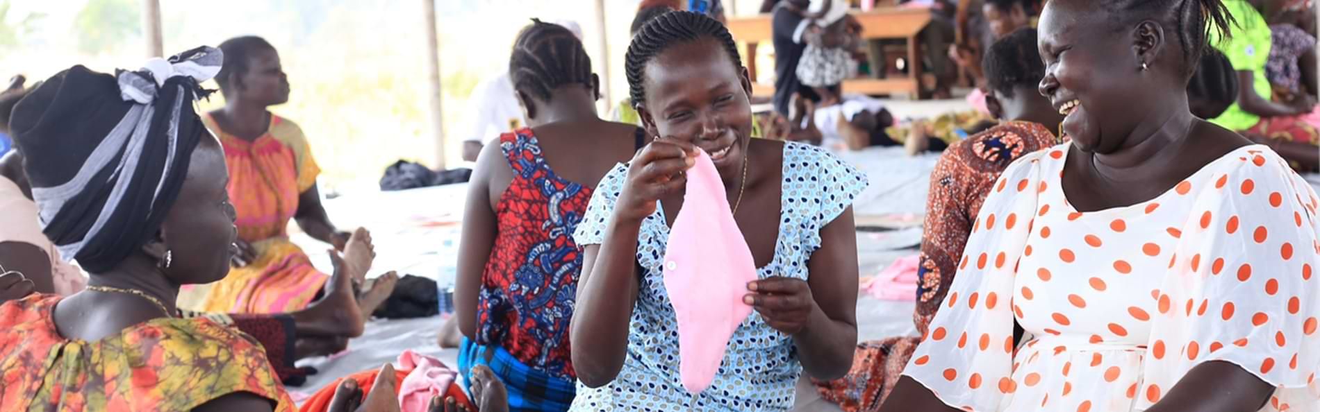 Eine Gruppe von Frauen in bunten Kleidern in einer Geflüchtetensiedlung in Uganda sieht sich eine Stoffbinde an und lacht.