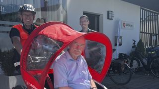 Mann in hellblauem Hemd wird in der roten Malteser Rikscha von einem Mann mit Fahrradhelm und oranger Weste gefahren, daneben steht ein weiterer Mann