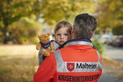 Ein Mann in Malteser Jacke trägt ein Kind mit Teddy auf dem Arm