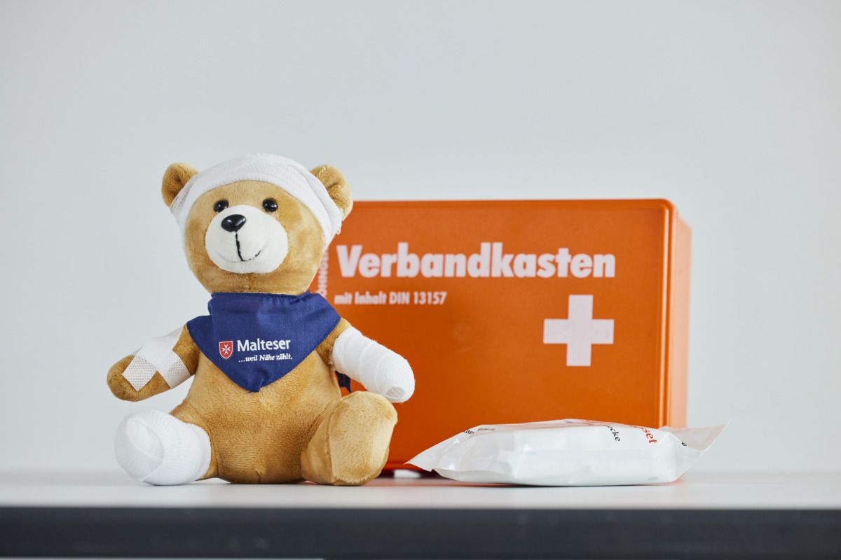 Teddybär mit blauem Malteser Halstuch und Verbänden an mehreren Körperteilen sitzt vor einem orangen Verbandkasten