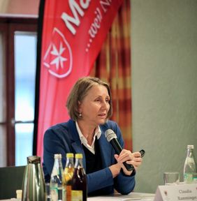 Frau mit Mikrofon bei Vortrag, sitzt am Tisch mit Getränken, roter Malteser-Banner im Hintergrund.