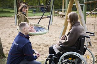 Ein Mann in einer blauen Malteser-Jacke hockt neben einer Frau in einem brauen Mantel, die im Rollstuhl sitzt. Beide sind auf einem Spielplatz. Im Hintergrund schaukelt ein Kleinkind und eine weitere Frau steht neben dem Kind und passt auf.