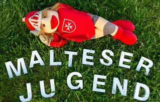 Handpuppe liegt über einem Schriftzug "Malteser Jugend" in der Wiese